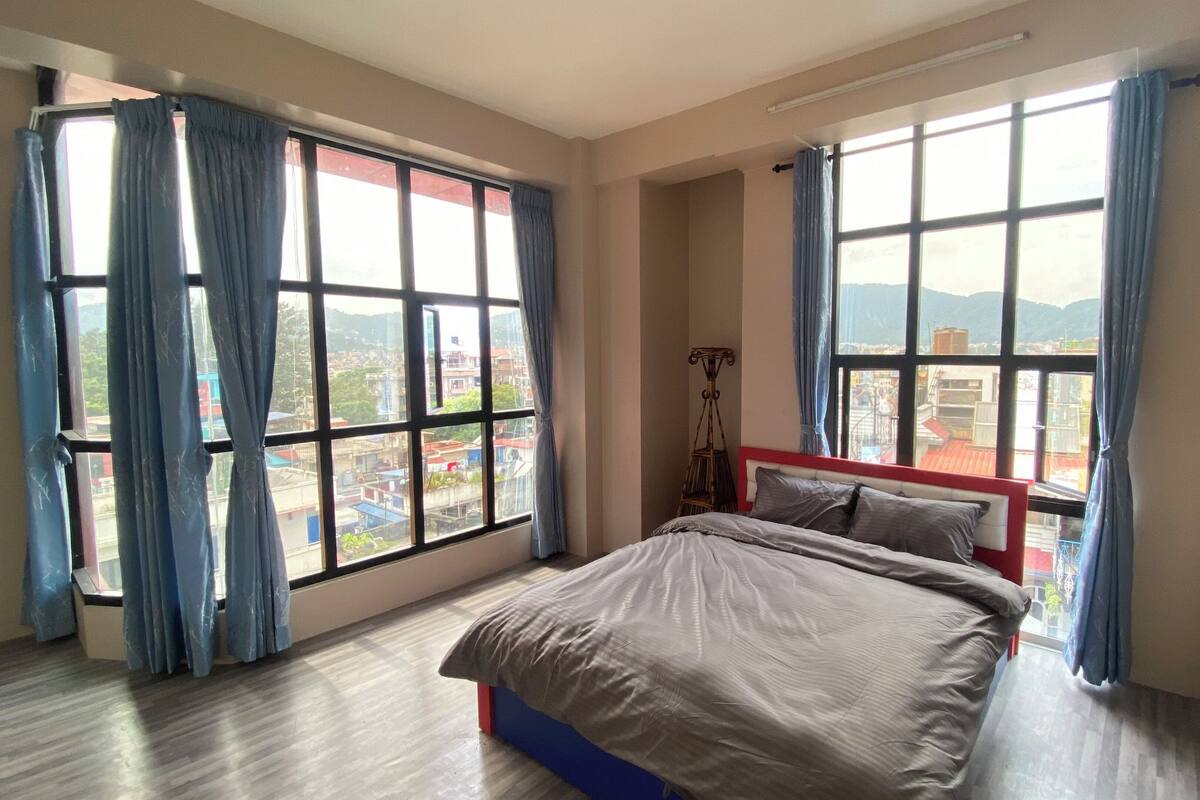 A bedroom with a bed, large windows, and a view of the city.