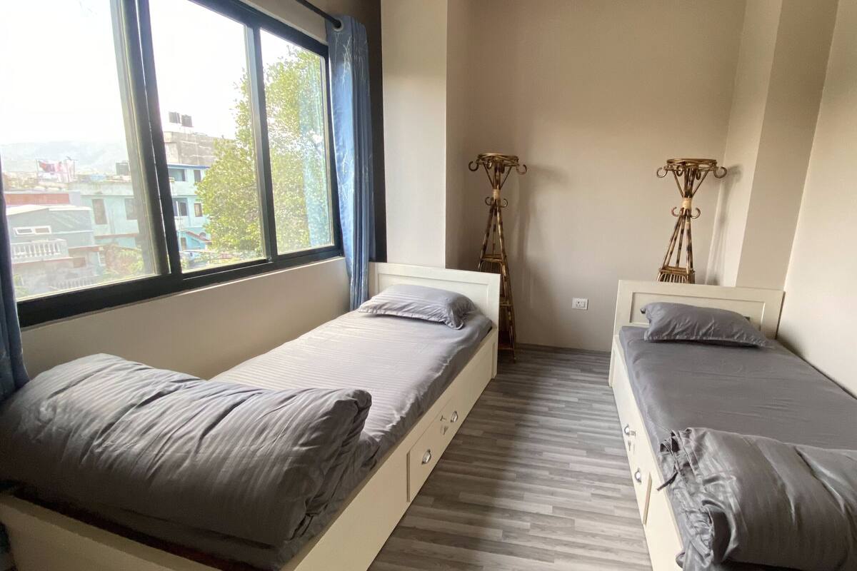 A small, clean room with two single beds, a window with curtains, and a view of buildings outside.