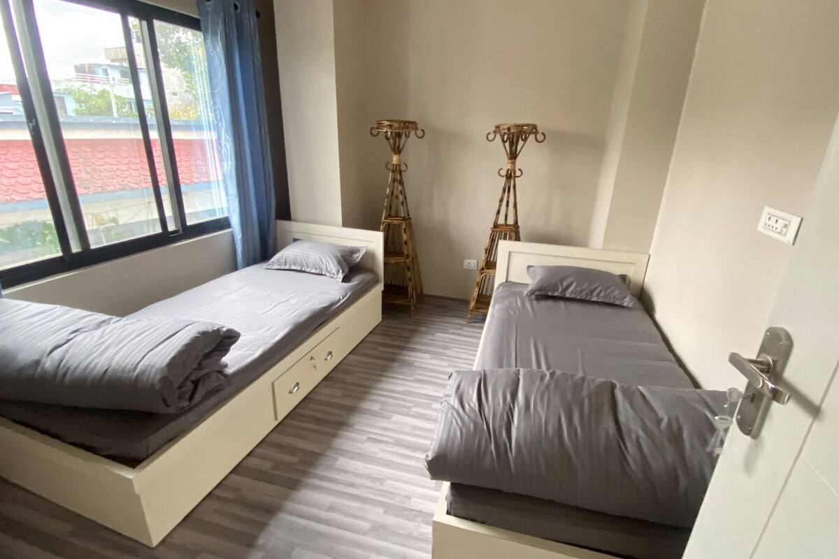A room with two single beds, a window with curtains, a small table, and a chair.