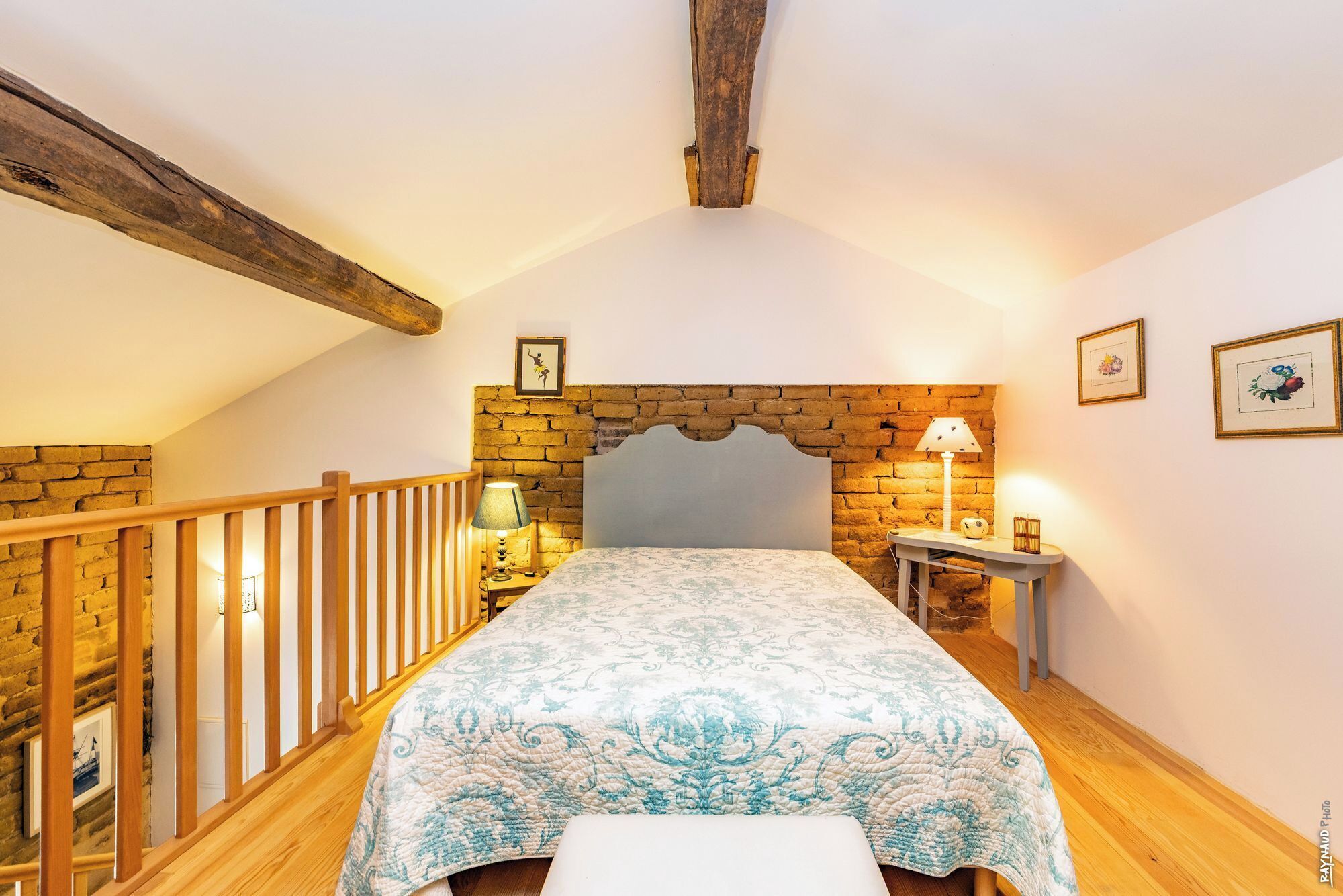 A bedroom with a stone wall headboard, a wooden railing, a bed with a patterned bedspread, a bedside lamp, and a small table with a lamp.