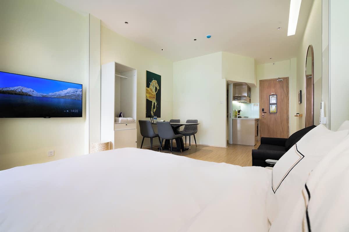 A hotel room with a bed, a TV mounted on the wall, a dining area with chairs and a table, and a kitchen area with a refrigerator and cabinets.