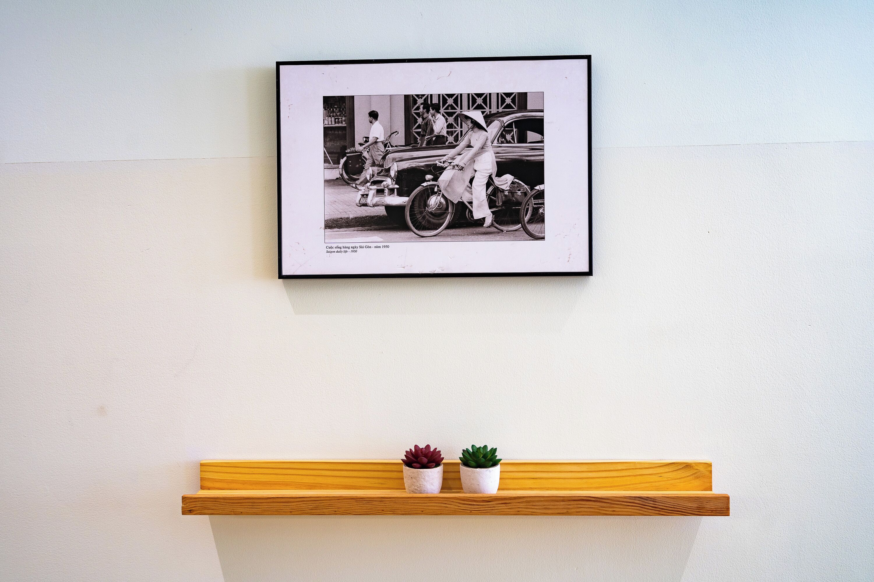 Fotografía en blanco y negro enmarcada que muestra un coche antiguo y una bicicleta sobre una pared, con un estante de madera en la parte inferior que sostiene dos macetas pequeñas.