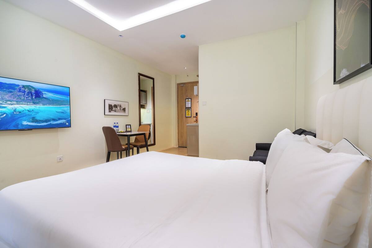 A hotel room with a bed, a desk with a chair, a TV on the wall, and a door leading to another room.