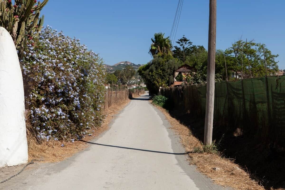 Una estrecha vía pavimentada bordeada por vegetación y una cerca, que conduce hacia un paisaje montañoso.