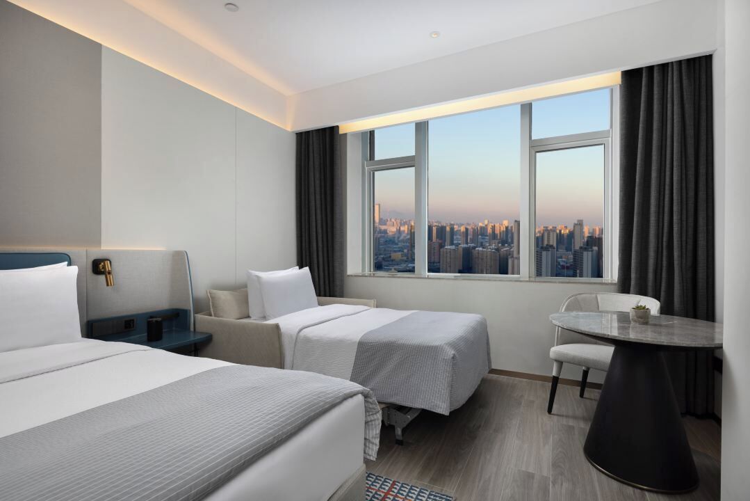 A hotel room with two beds, a small table, a chair, and a view of a cityscape.