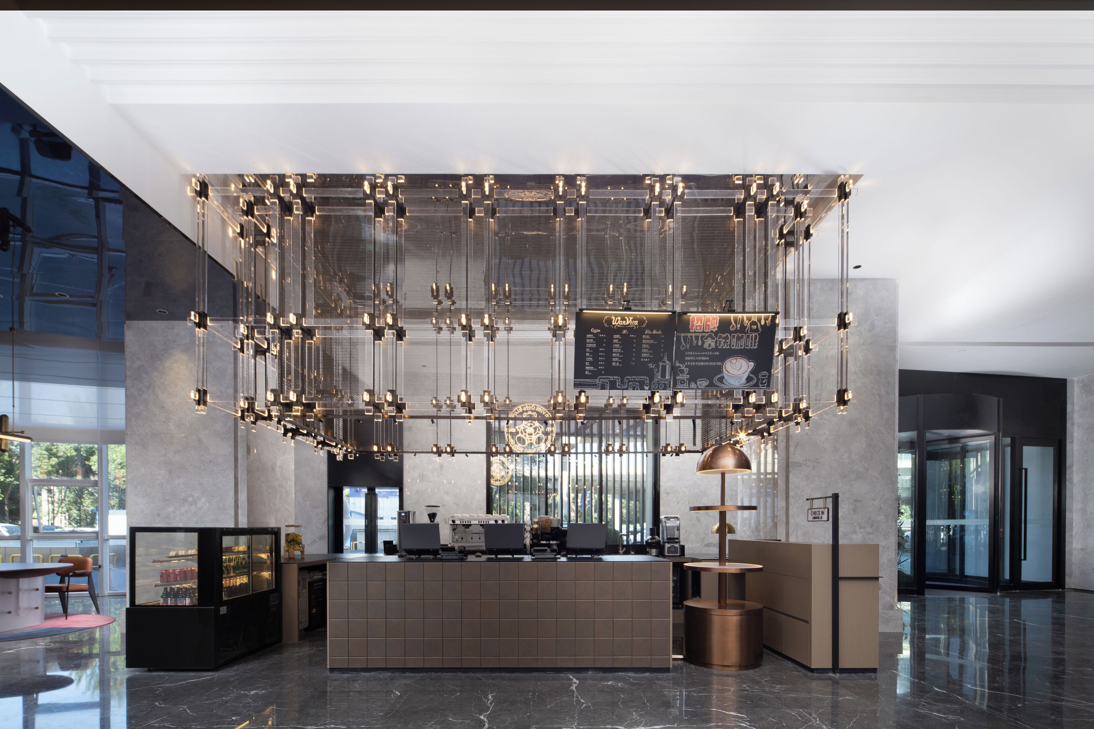 A modern hotel lobby with a central reception desk, a large chandelier, and a glass partition.