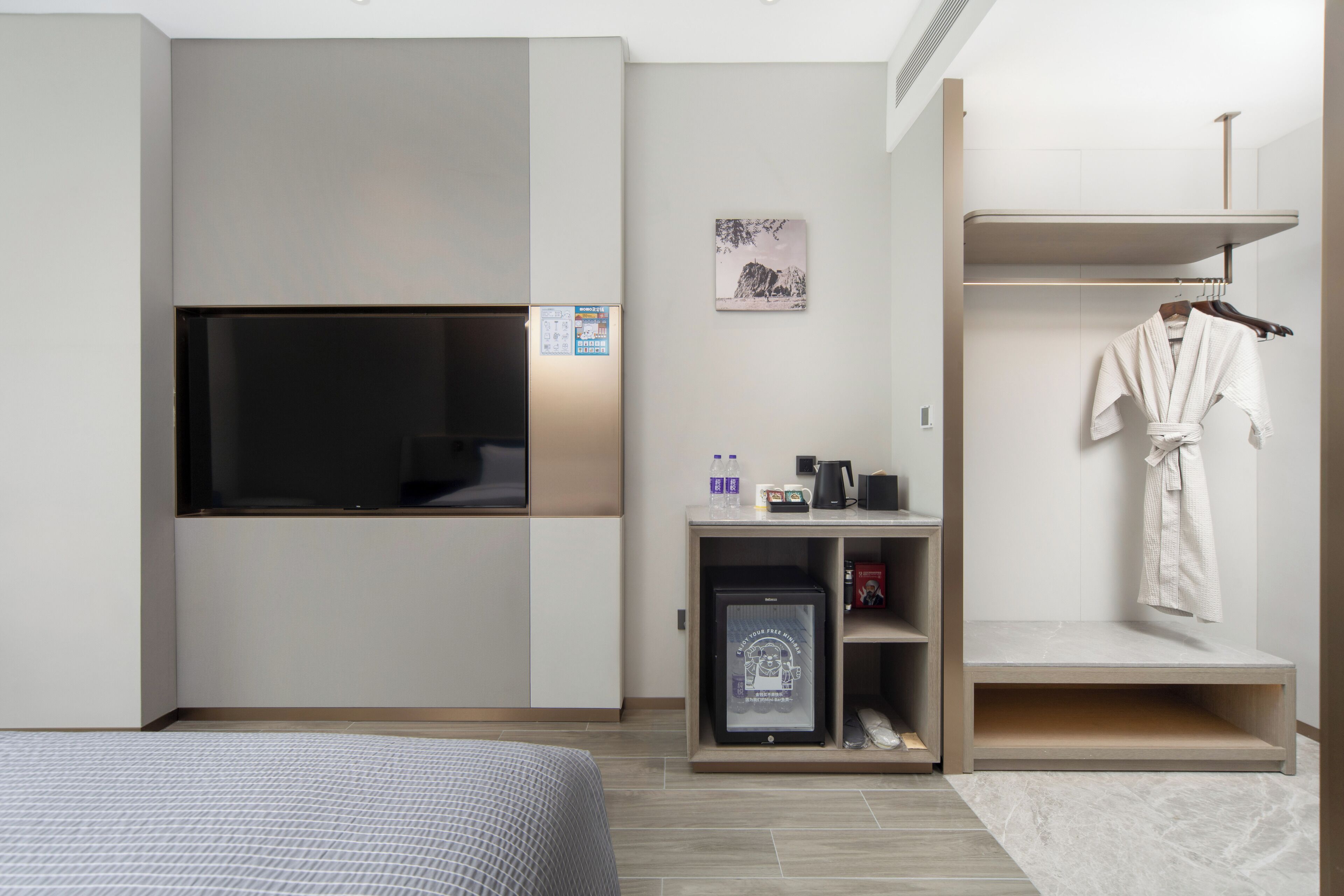 A modern hotel room with a bed, a TV mounted on the wall, a small fridge, a shelf with toiletries, and a robe hanging on a rack.