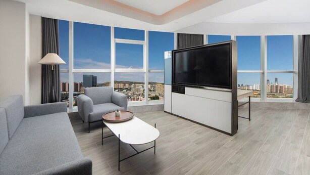 A modern hotel room with a large TV, grey sofa, a small table, and a city view.