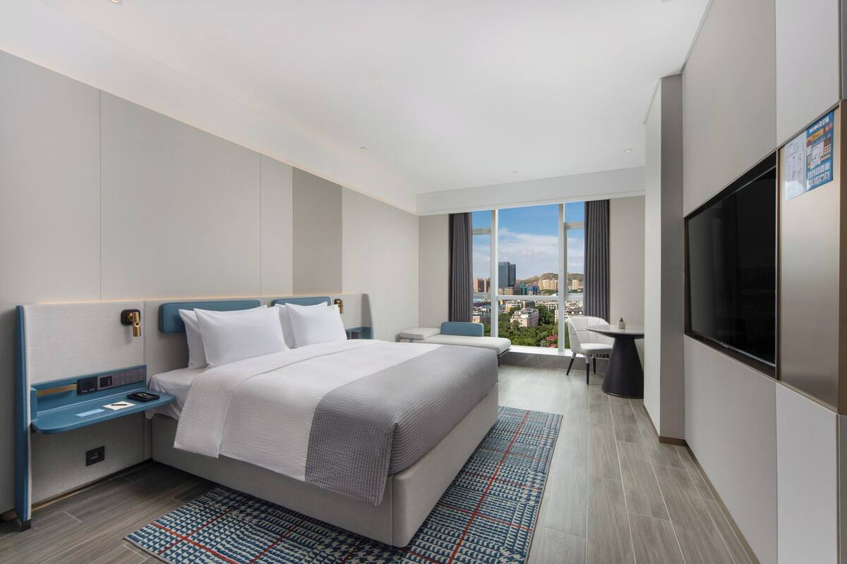 A modern hotel room with a large bed, a desk, a TV, and a view of the city.