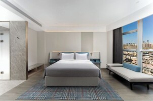 A modern hotel room with a large bed, bedside tables, a sofa, and a city view.