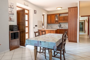 A dining area with a table set for four, wooden cabinets, a kitchen in the background, and a door leading to another room.