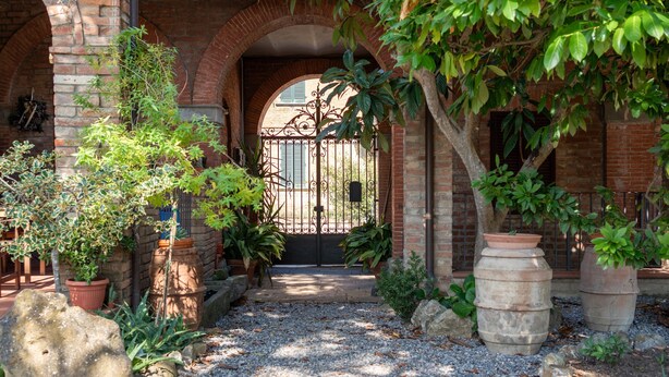 Un chemin de jardin mène à un bâtiment en briques, surmonté d’un portail en métal, et est bordé de plantes en pot et de rochers.