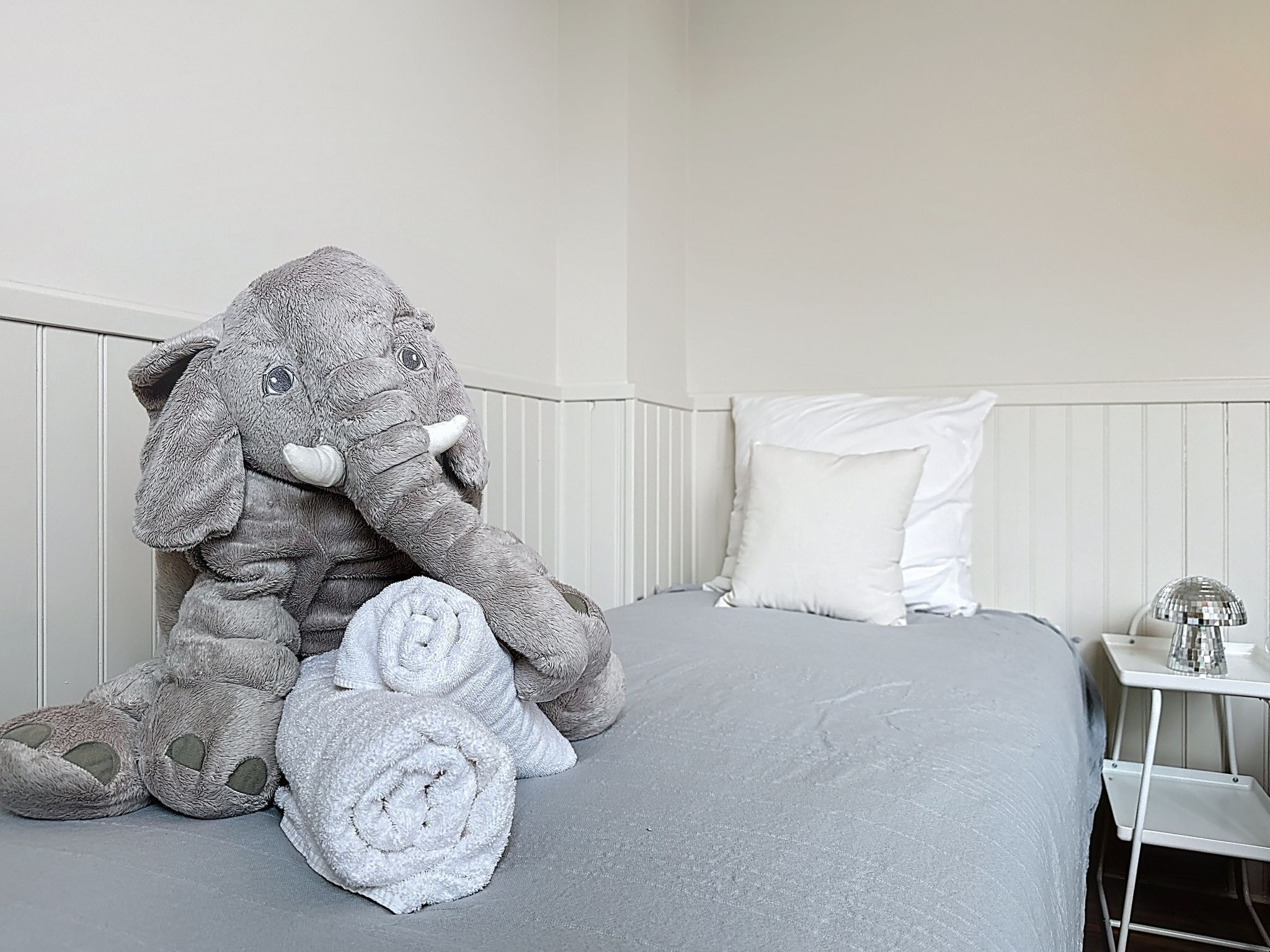 Een net opgemaakt bed met een grijze deken, een wit kussen en een grote grijze knuffelelefant.