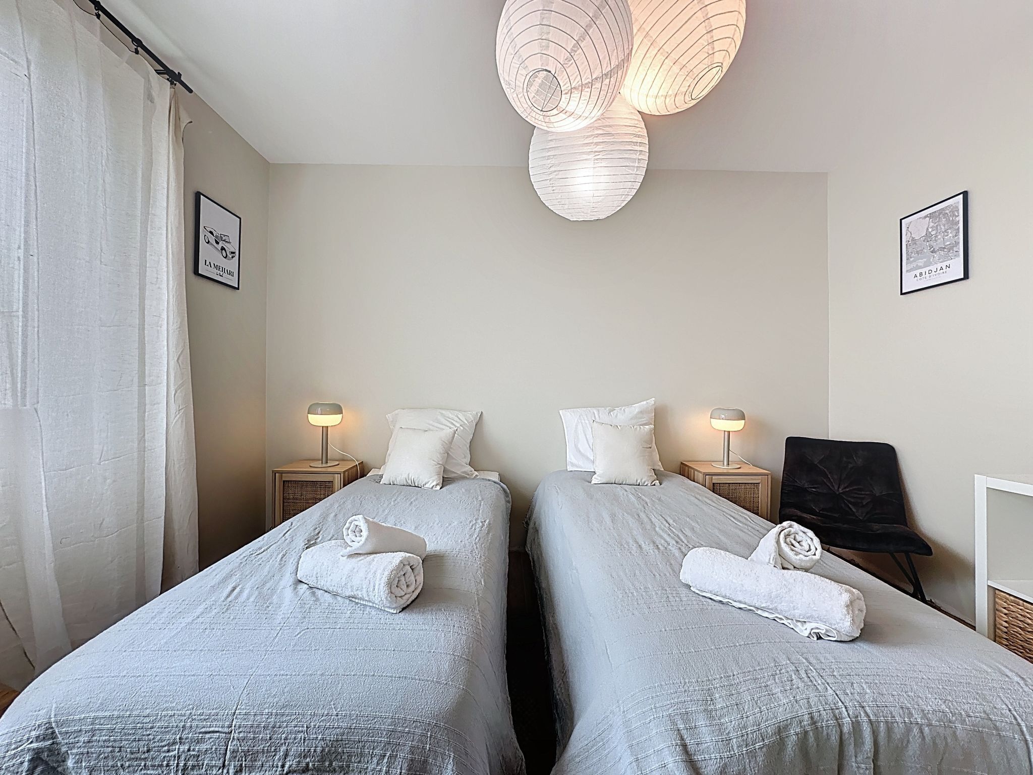 Hotelkamer met twee bedden, elk met een wit kussen en een opgerolde handdoek. Er staan nachtkastjes met lampen, een zwarte stoel, en er hangen twee ingelijste foto's aan de muur.