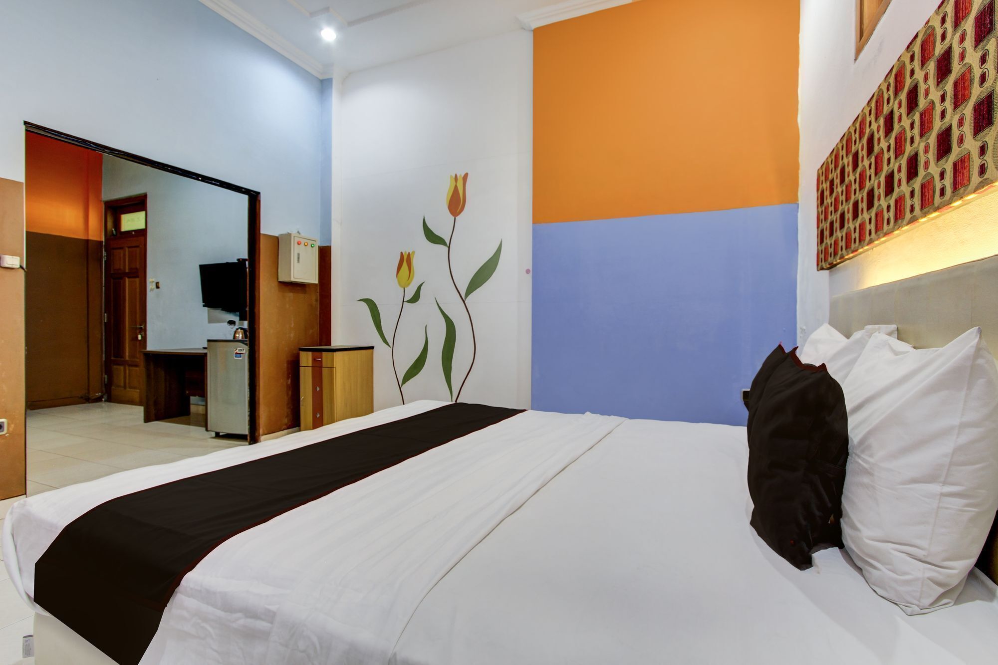 A hotel room with a bed, a desk, a chair, a television, and a wall with a floral mural.