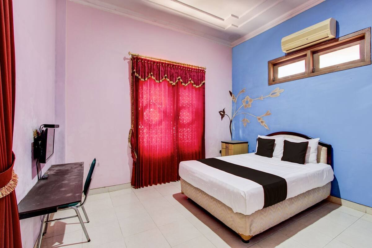 A bedroom with a bed, a desk, a chair, a window with curtains, and an air conditioning unit.