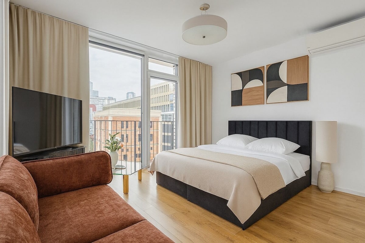 A modern hotel room with a large bed, a flat-screen TV, a sofa, and a city view through large windows.