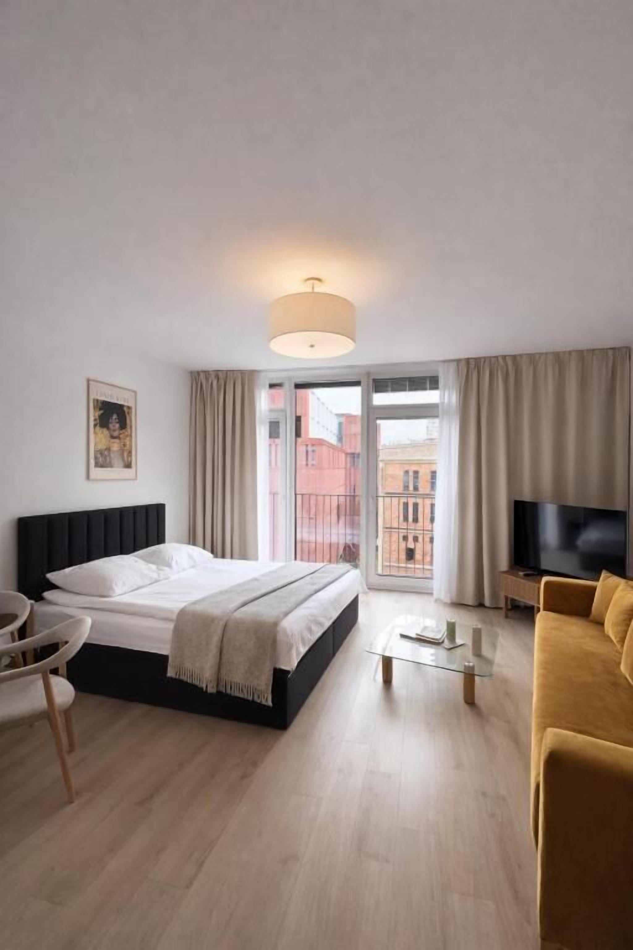 Ein Hotelzimmer mit Bett, Fernseher, Sofa, Sessel und einem Balkon mit Blick auf Gebäude.