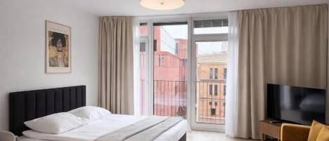 Habitación de hotel con cama, televisor, sofá, sillón y balcón con vista a edificios.