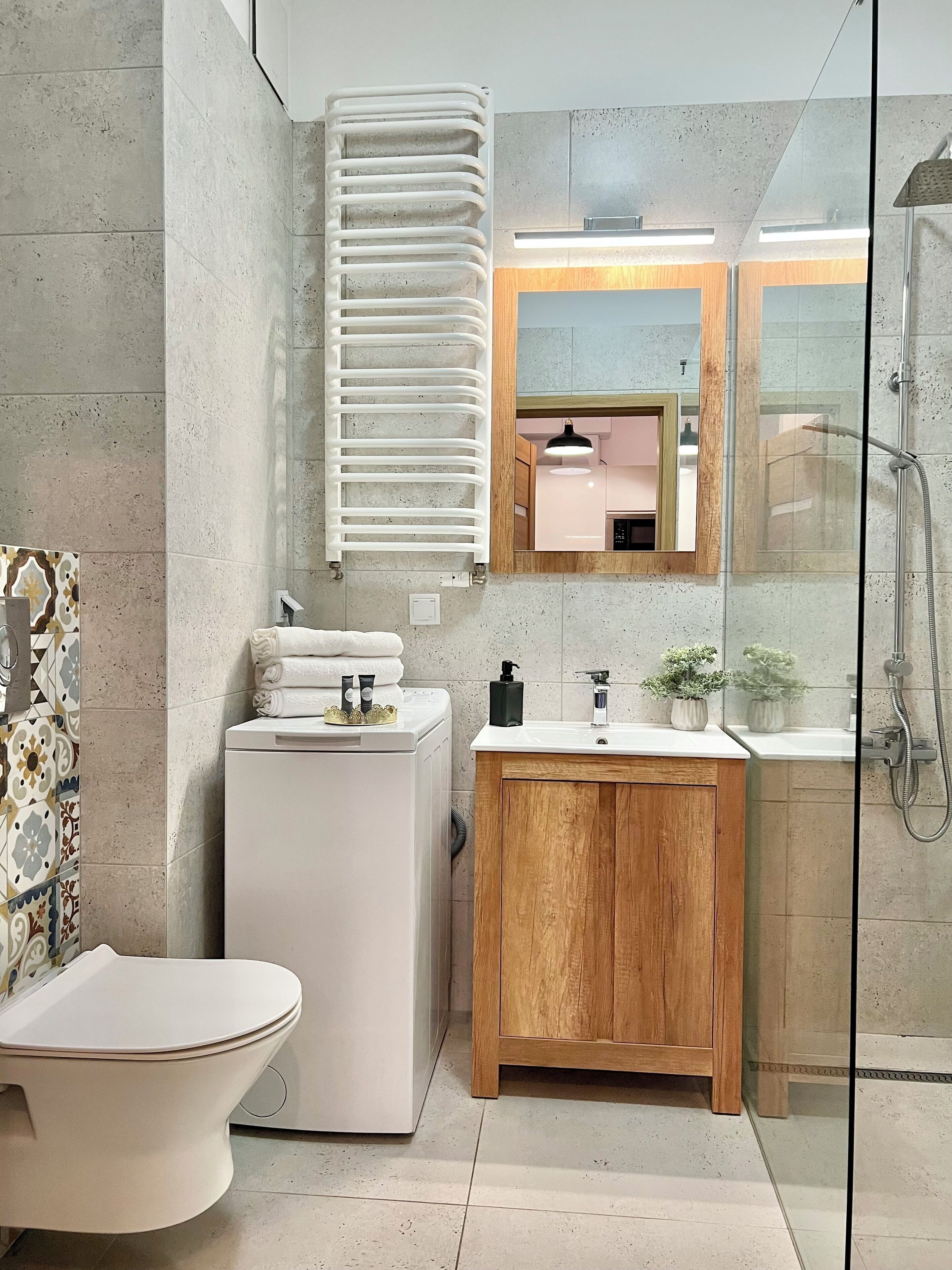 A modern bathroom with a freestanding toilet, a built-in washing machine, a wooden vanity with a sink, and a glass-enclosed shower area.