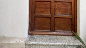 A wooden door with a metal handle, a sign reading "QORI SICILIAN STAY" above it, and a wall-mounted light fixture to the left.