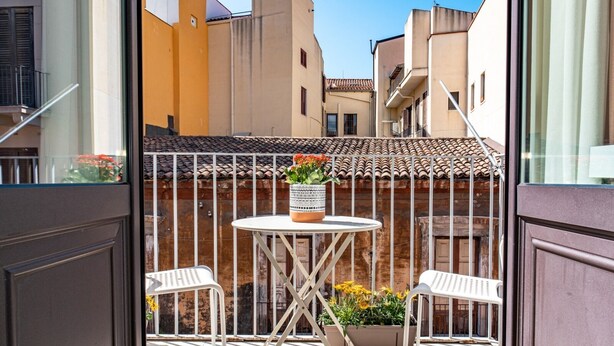 A balcony with a round table and two white chairs, a potted plant with flowers, and a view of neighboring buildings.