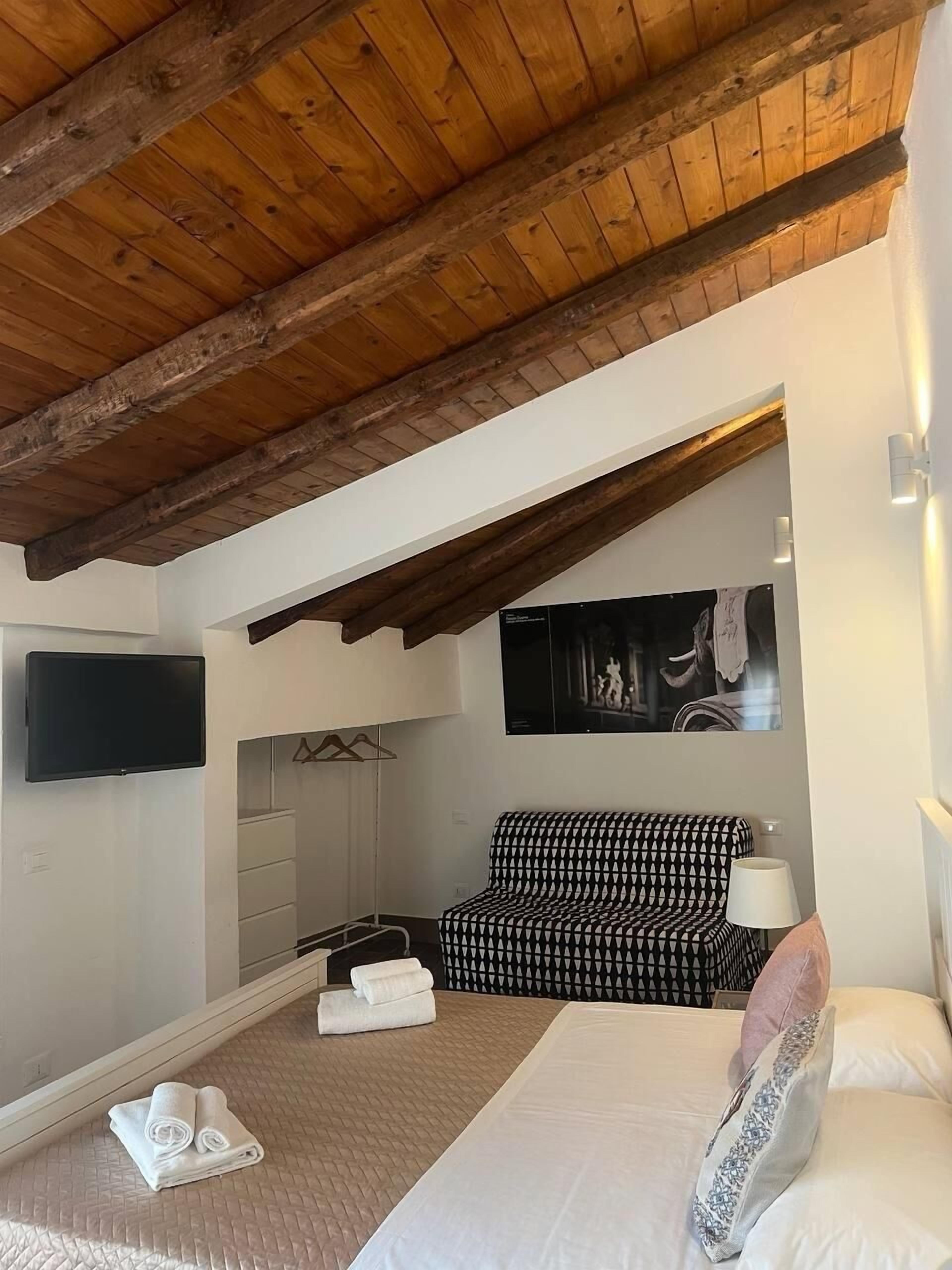 Een slaapkamer met een houten plafond, een bed met wit beddengoed, een gestreept sofa, een flatscreen televisie en een kunstwerk aan de muur.