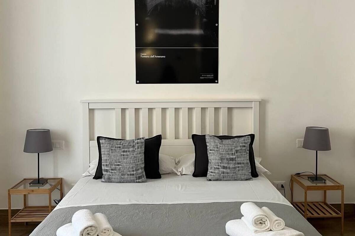 A hotel room with a bed, two nightstands, a night lamp on each, and a poster of a fountain on the wall.
