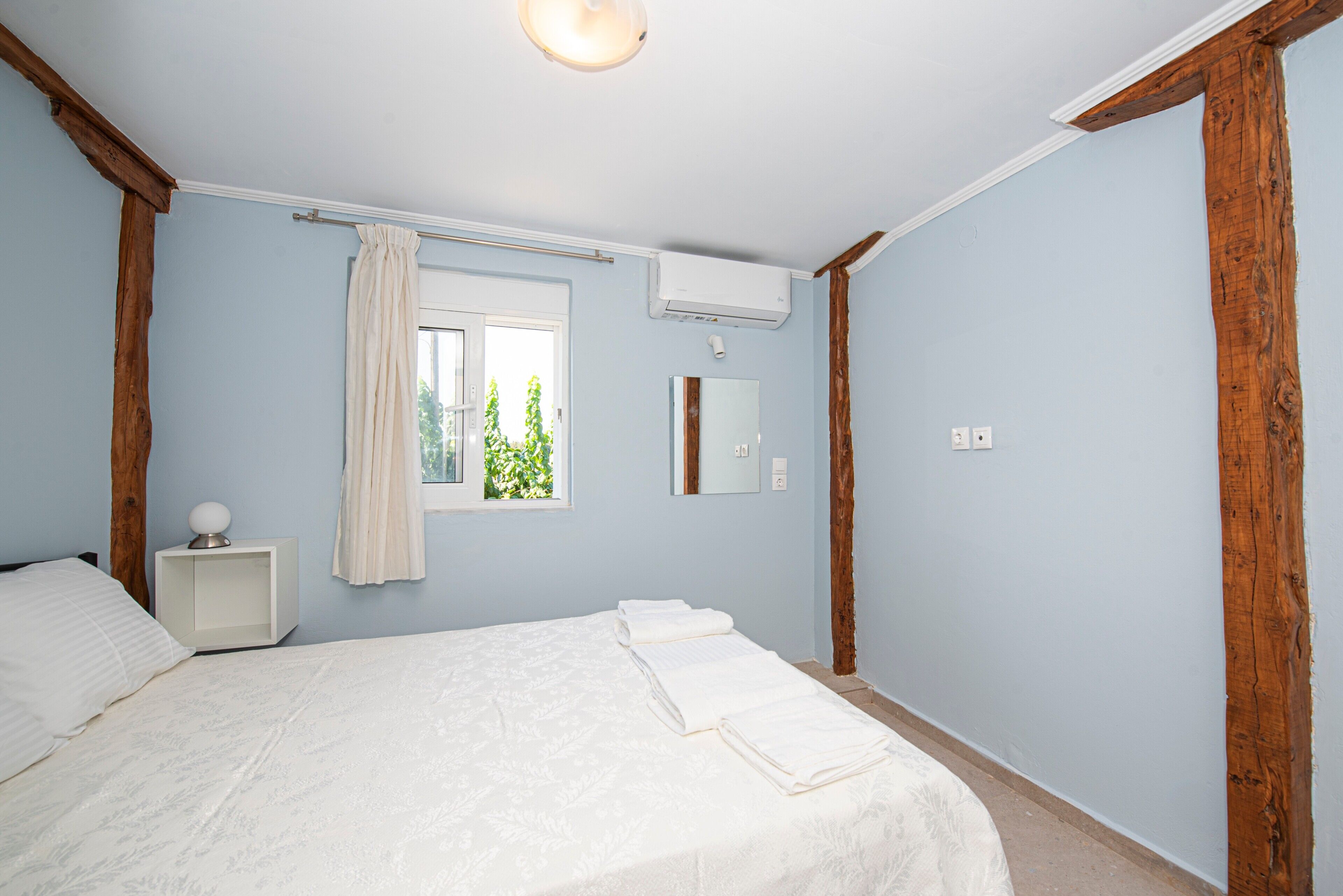A bedroom with a bed, a nightstand, a window with curtains, a mirror, and an air conditioning unit.