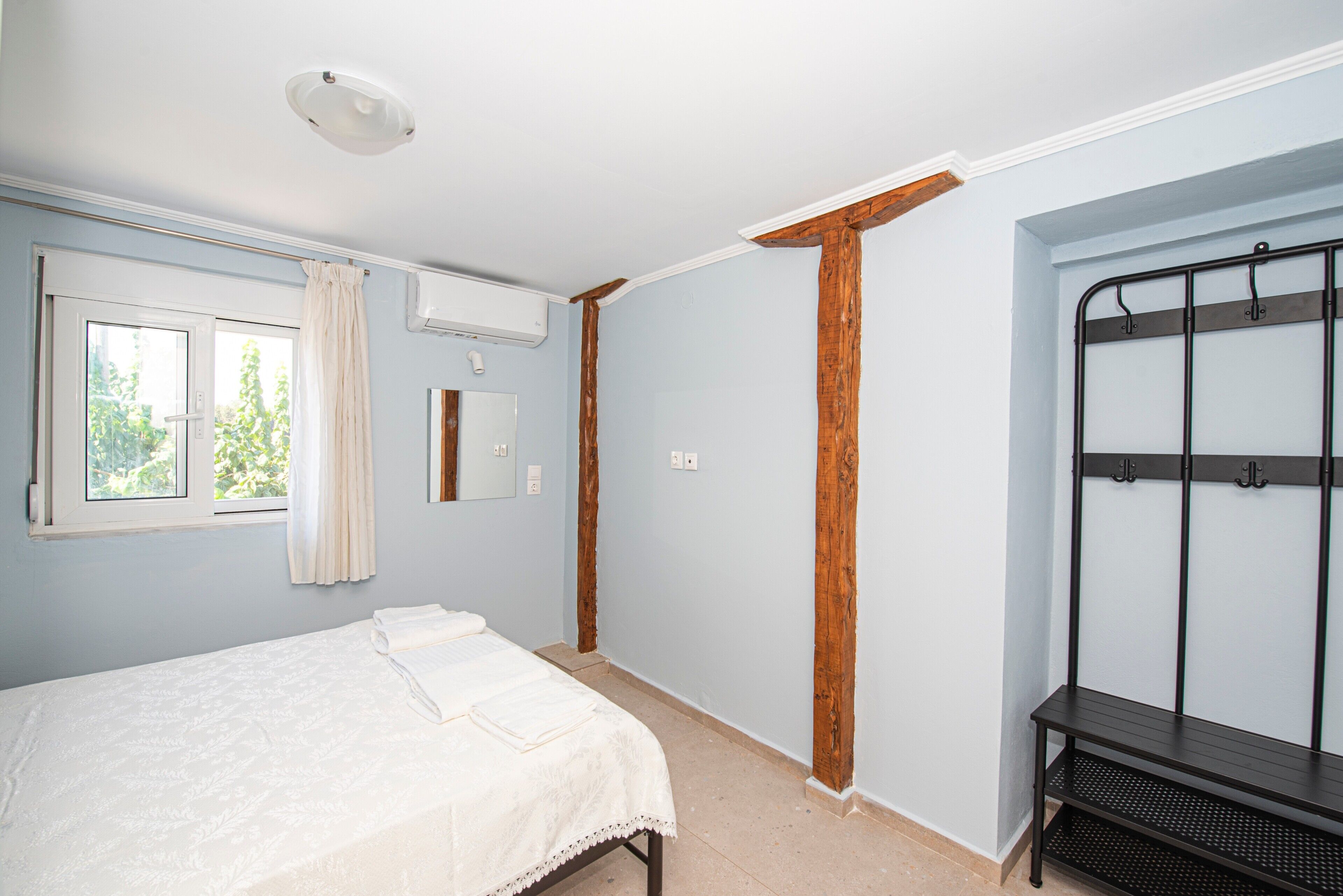 A bedroom with a bed, a window with curtains, a wall-mounted air conditioner, and a metal bench.