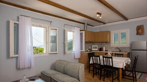 A cozy living room with a sofa, a glass-top coffee table, and a dining area with a table set for four. A kitchen with wooden cabinets and a microwave is visible in the background. - Holiday Home 'Lovely' with Balcony, Wi-Fi and Air Conditioning (Sivros)