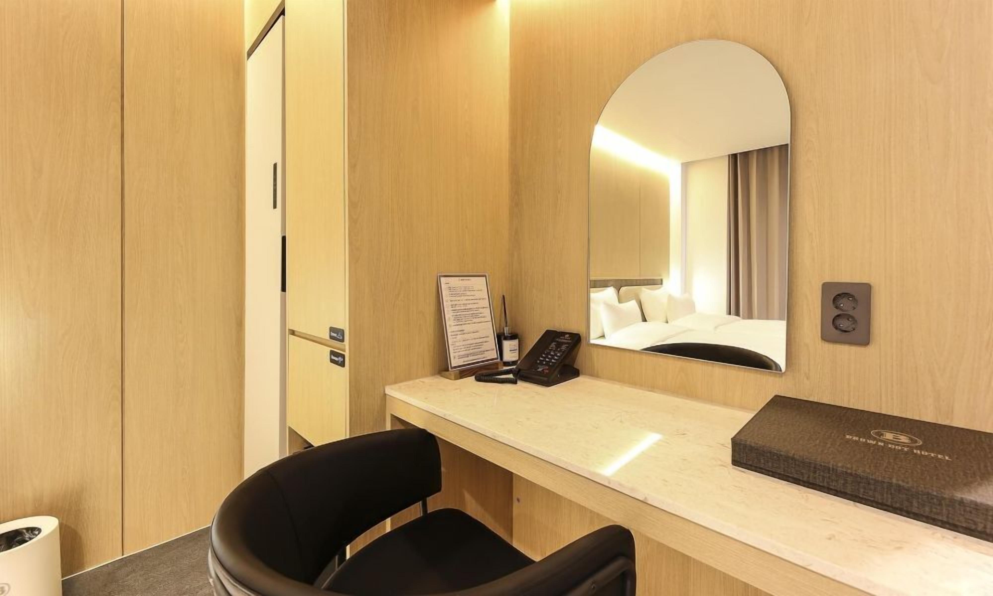 A hotel room with a wooden desk, a chair, a bed, a mirror, a bedside table, and a sign.