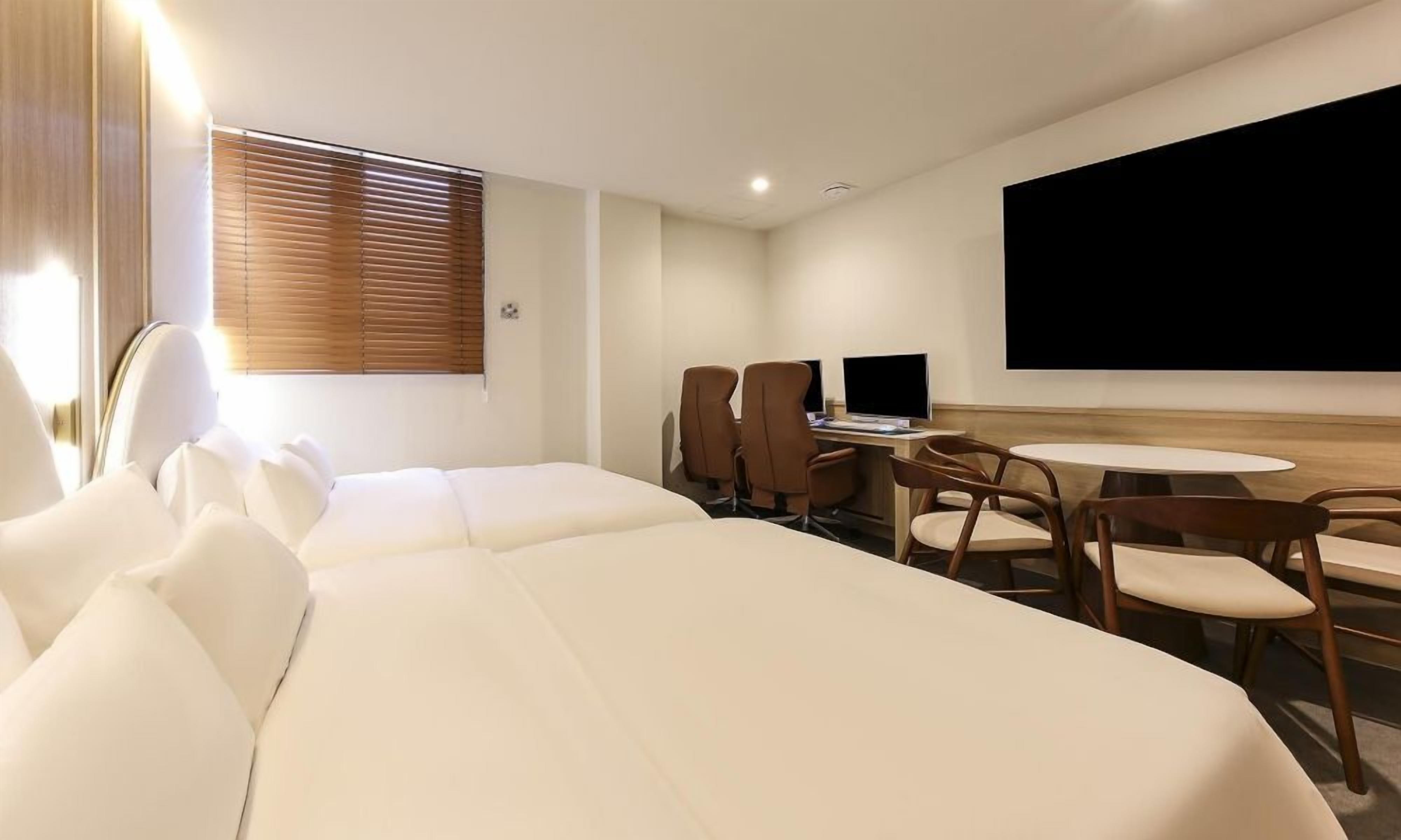 A hotel room with two beds, a desk with a computer, and a large TV screen.