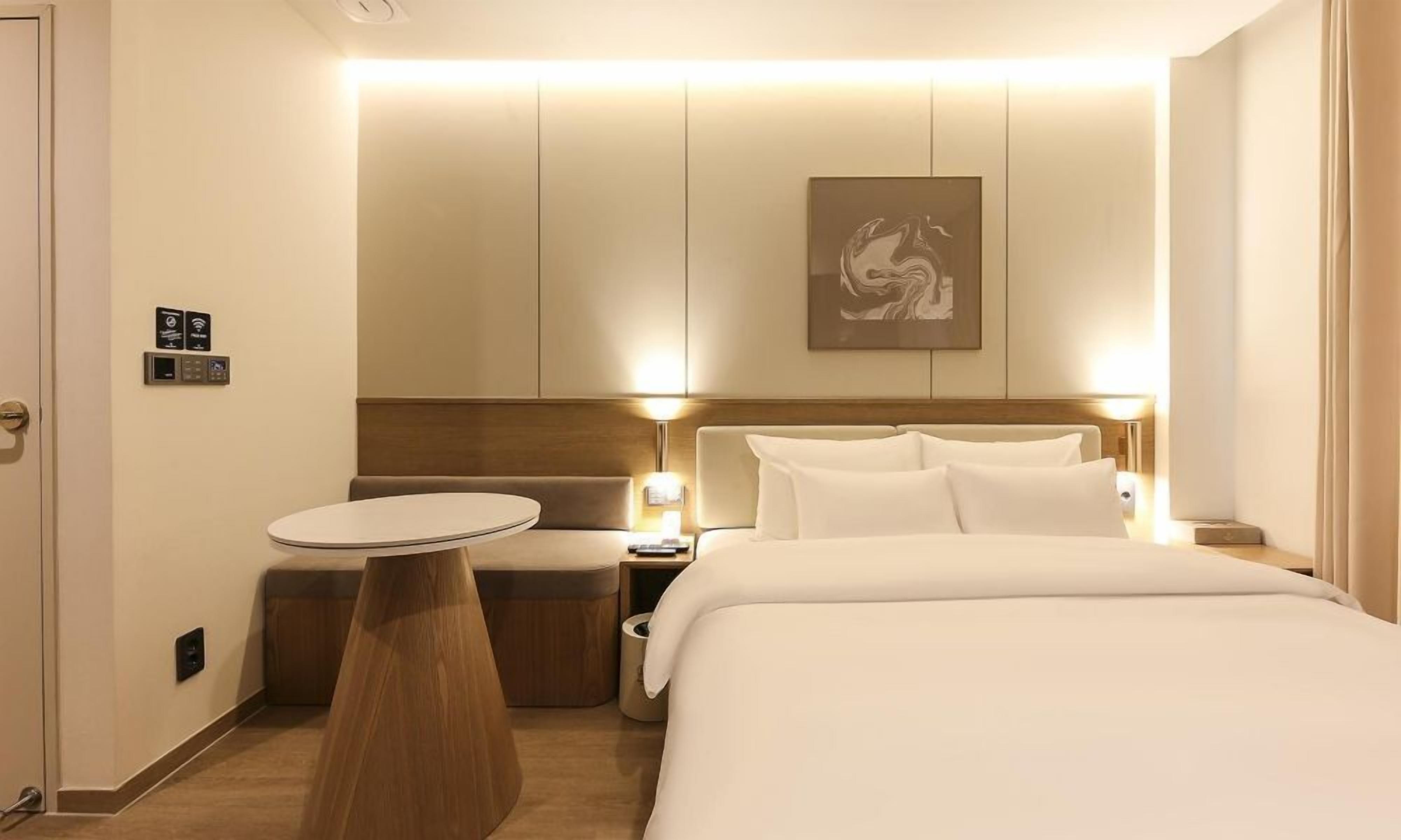 A hotel room with a bed, bedside table, and a small round table. There is a wall-mounted artwork above the bed.