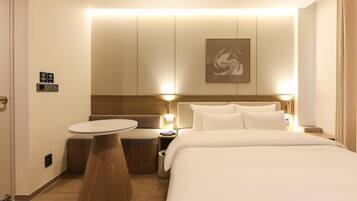 A hotel room with a bed, bedside table, and a small round table. There is a wall-mounted artwork above the bed.