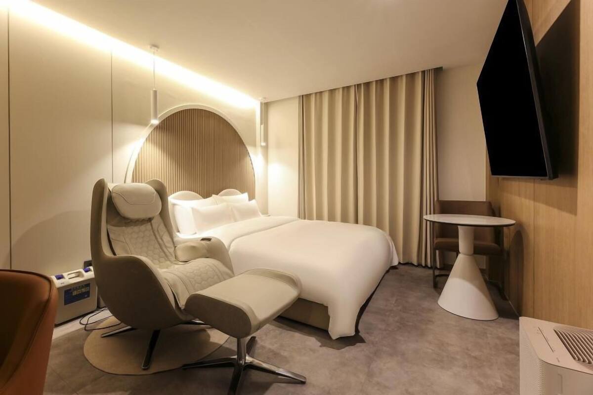 Ett modernt hotellrum med en stor säng, en sittgrupp med en fåtölj och ett litet bord, samt ett runt soffbord och en tv som är monterad på väggen.