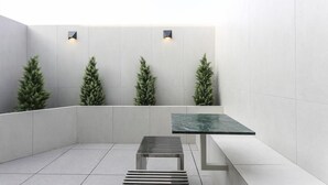 Quarto moderno e minimalista com piso ladrilhado, uma mesa central e três árvores de Natal artificiais em frente a uma parede lisa.