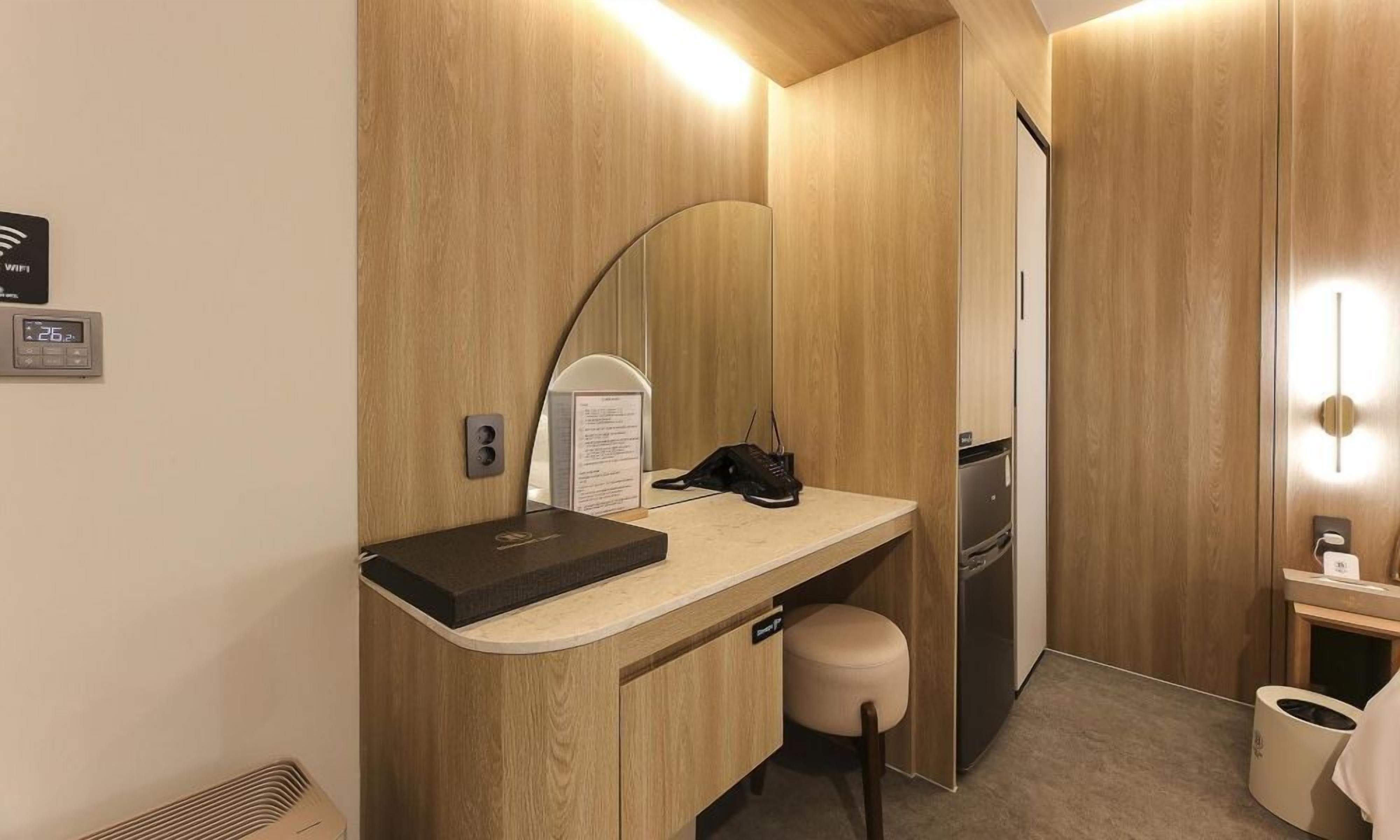 A compact hotel room with a wooden vanity, a mirror, a desk with a phone, a small refrigerator, and a trash bin.