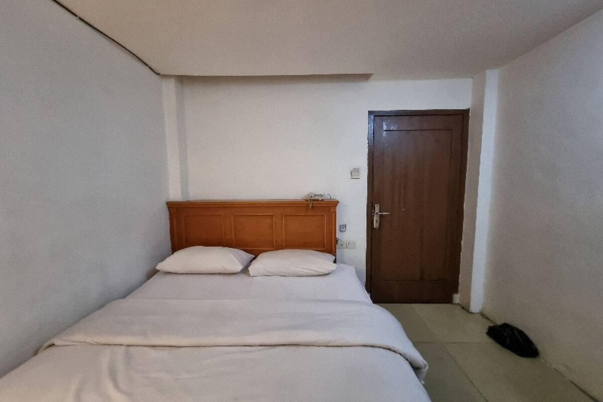 Una habitación pequeña y sencilla con una cama individual, cabecero de madera, una puerta y una bolsa pequeña en el suelo.