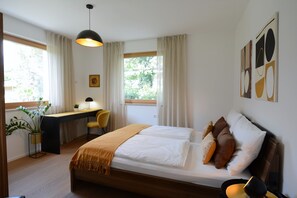 Quarto com cama, mesa de cabeceira, luminária, escrivaninha, cadeira e janela com cortinas.