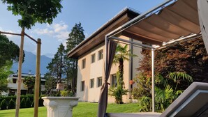 Zona relax esterna moderna con una sedia in metallo e un tavolo, immersa in un giardino curato con fontana, alberi e un edificio sullo sfondo.