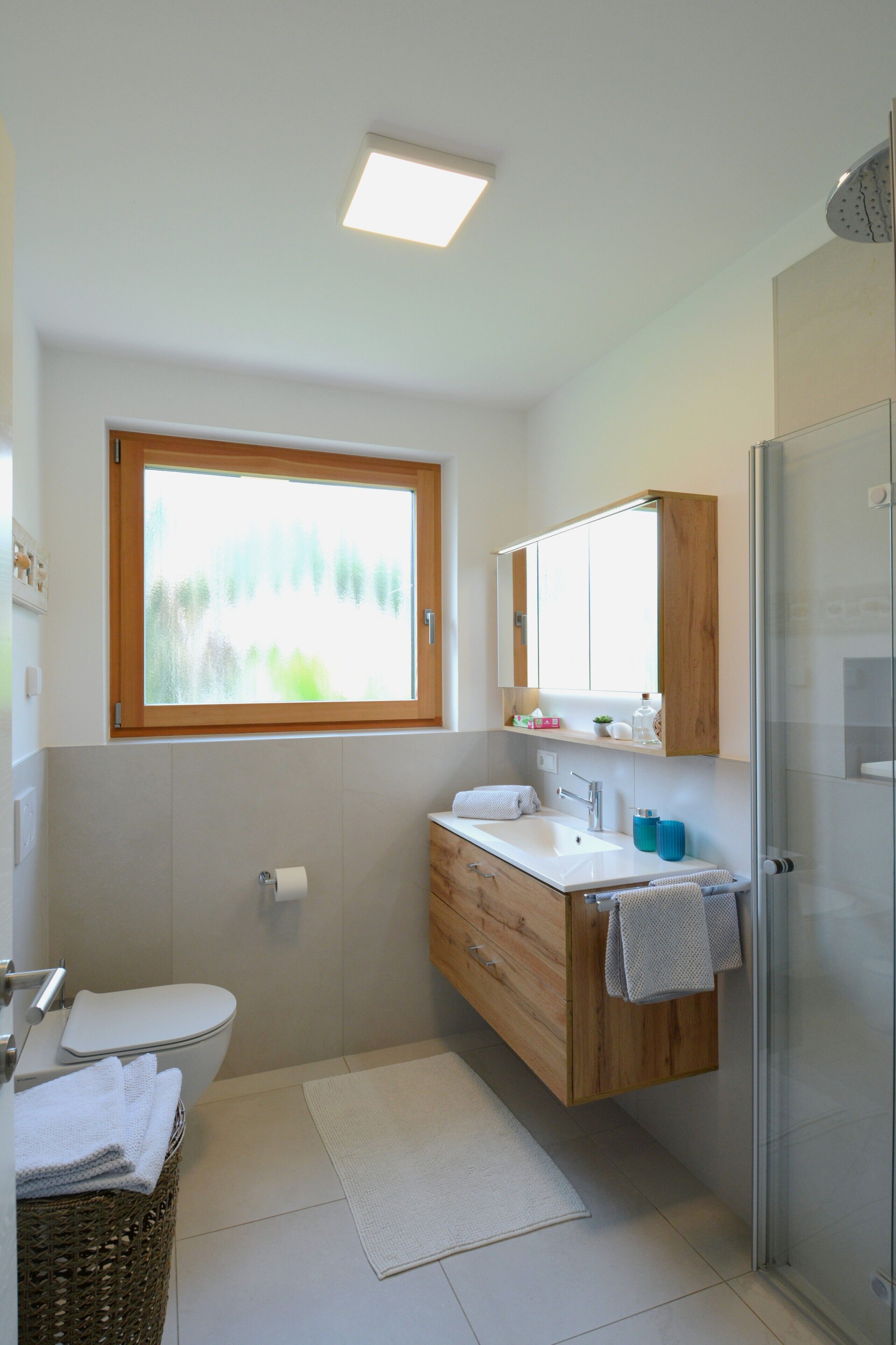 A modern bathroom with a wooden vanity, a glass shower enclosure, and a window providing natural light.