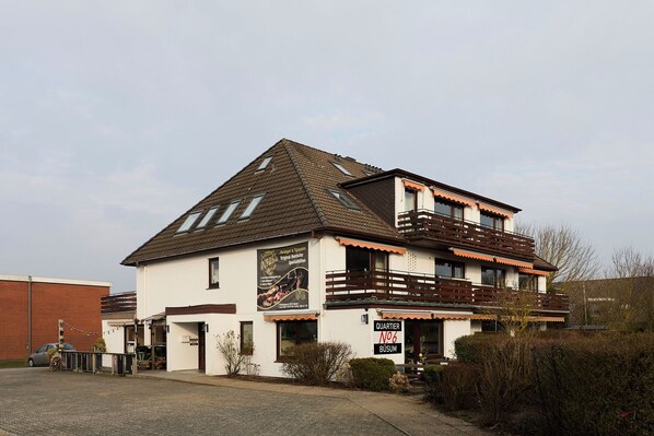 A two-story building with a balcony, a sign indicating it's a business, and a small garden area in front. - Quartier 3 apartment in a quiet location and access to the sea time (Büsum)