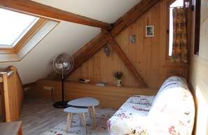 A cozy attic room with a sloped ceiling, wooden beams, a fan, a small table, a sofa, and a window with a curtain.
