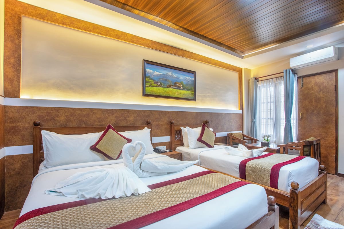 Una habitación de hotel con una cama grande, dos camas más pequeñas, cabeceros de madera, un cuadro de un paisaje montañoso y una ventana con cortinas.