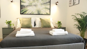 A neatly made bed with folded towels, a large abstract painting above it, and two potted plants on either side.