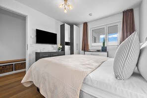 A modern bedroom with a large bed, a flat-screen TV, a wardrobe, and a window with curtains. - Enjoy - Parking space, balcony, grill, washing machine, first row on the water (Olpenitz)