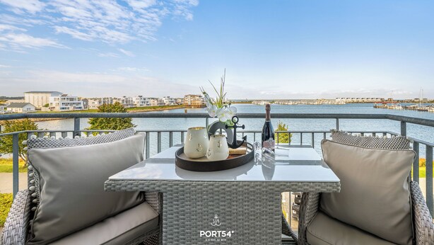 A balcony with wicker furniture, a table with a bottle and glasses, and a view of a marina and buildings across the water.