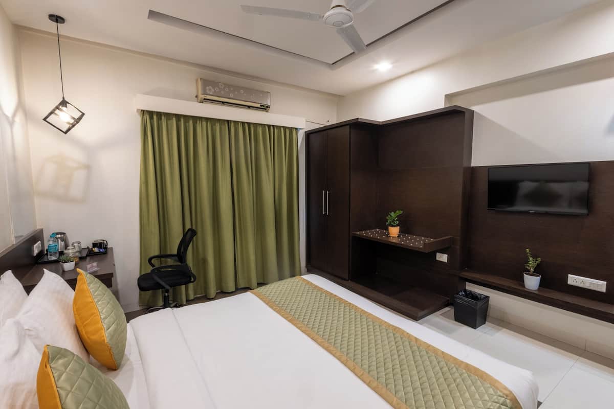 A hotel room with a bed, a desk with a chair, a TV mounted on the wall, a ceiling fan, and a window with curtains.