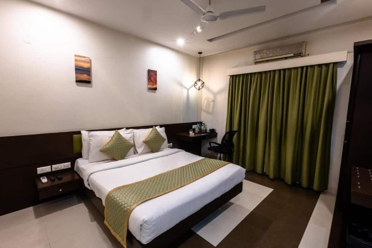 A hotel room with a large bed, a desk, a chair, a nightstand, a wall-mounted air conditioner, and a window with green curtains.